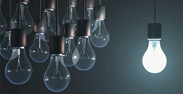 An illuminated light bulb shines by itself as eight unlit bulbs surround it.