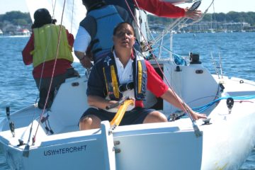 Wearing a red, white and blue shirt under his lifejacket, a blind sailor steers a sailboat.