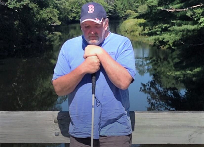 Deon Lyons stands on a bridge over a small river holding his cane. Hand over hand on top of his cane, he rests his chin on top of it while deep in thought.