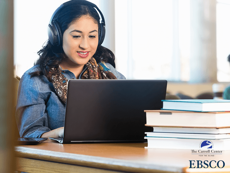 A woman wearing headphones smiles as she uses her laptop in the library. A stack of books rests on the desk.