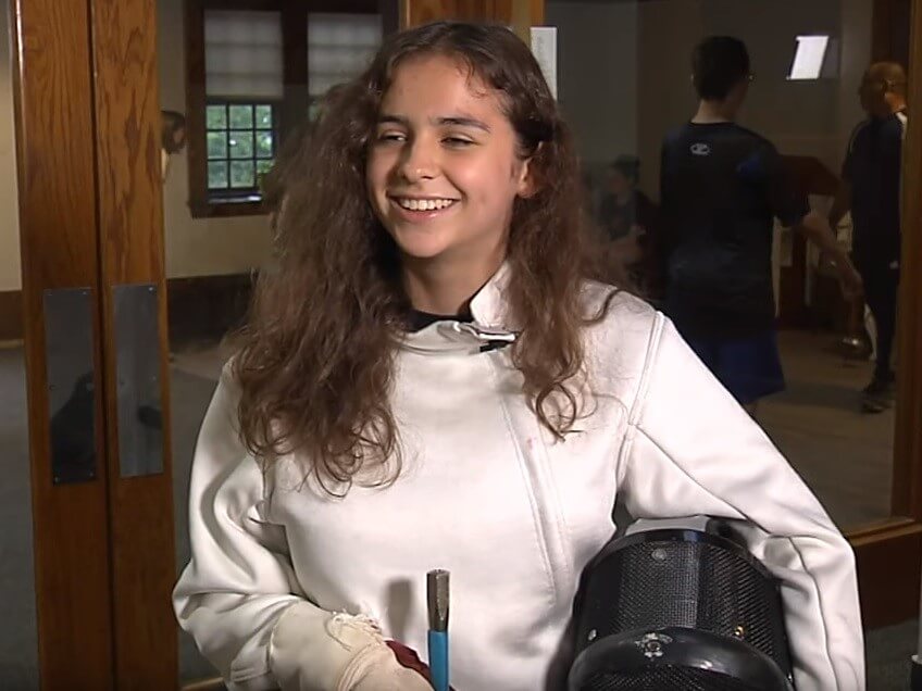 A female Carroll center summer student wears full fencing equipment during a media interview.