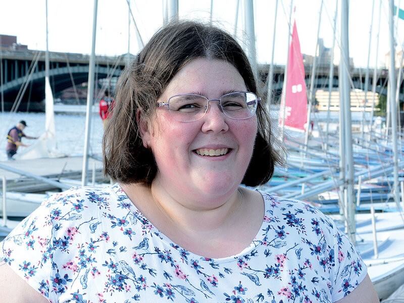 Katherine Kern, whose sailing origins began with the SailBlind program, smiles on a dock in front of a row of sailboats.