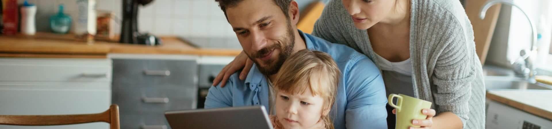 In a kitchen, a mother, father and young daughter huddle around a laptop.