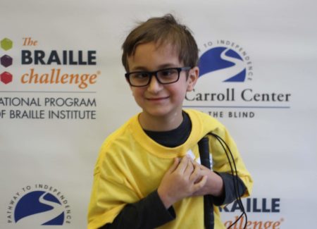Apprentice group winner, Benjamin Silva, wears a yellow braille challenge shirt as he poses in front of a braille challenge banner.