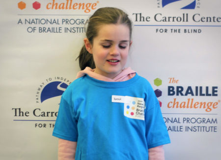 Freshman group winner, Hannah Gevers, wears a blue braille challenge shirt and poses in front of a braille challenge banner.