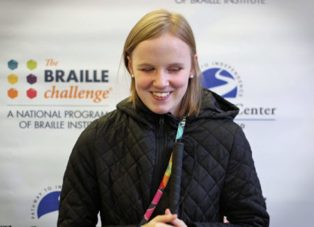 Varsity group winner, Paige Drury, smiles in front of a banner with the braille challenge and carroll center logos.