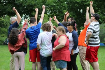 A group of over a dozen students huddle together and raise their fists in the air at the conclusion of an activity on the lawn.