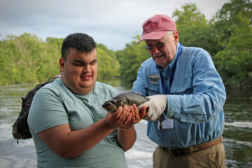 After catching a large green fish, David smiles wide as he holds it up in his hands.