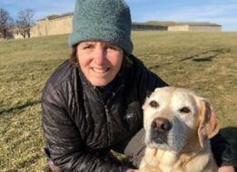 Martha Steele kneels beside her guide dog Alvin on a hill.