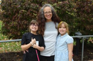 A mom with her daughters, Cane Quest participants, standing and smiling on the Carroll Center for the Blind campus outside