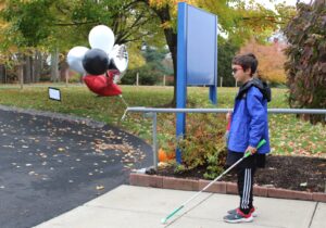 Cane Quest participant standing with his white cane on the Carroll Center for the Blind campus outside next to balloons