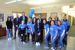 A group of corporate volunteers in blue T-shirts smiling for a photo with Greg Donnelly, Carroll Center for the Blind President and CEO. They are standing inside one of the Carroll Center campus buildings and have blue balloons behind them.