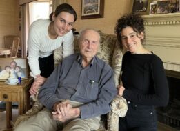 John sitting on a chair at his home with his grandaughters Eva and Anna standing next to him