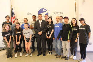 A group of corporate volunteers from Deloitte smiling for a group picture with Carroll Center President & CEO Greg Donnelly in front of The Carroll Center for the Blind logo wall