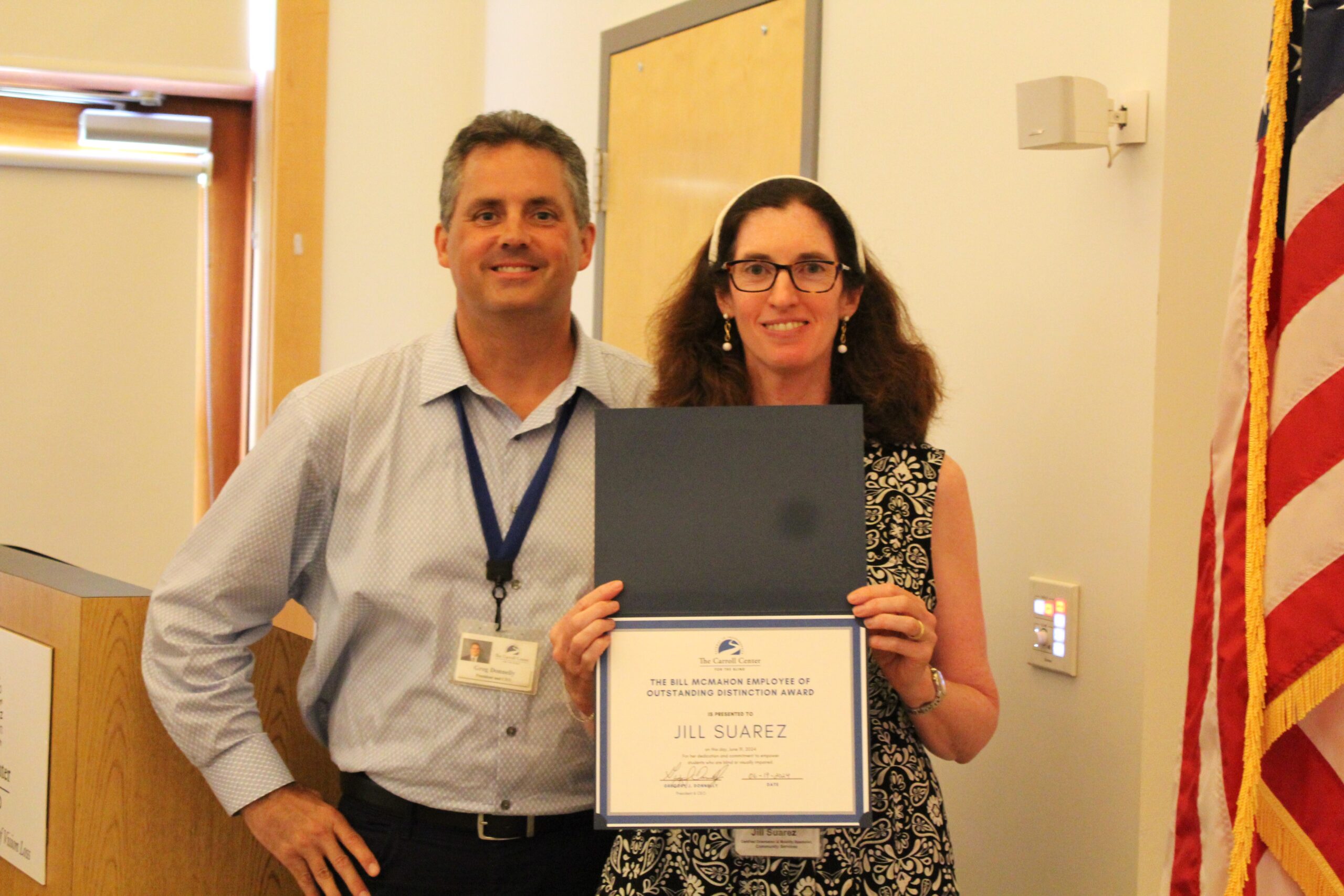 Jill Suarez and Carroll Center President & CEO, Greg Donnelly, stand together smiling in a brightly lit room with the American flag visible to the right. Jill is holding a blue certificate folder with an award. The text on the certificate reads: "The Bill McMahon Employee of Outstanding Distinction Award is presented to Jill Suarez."